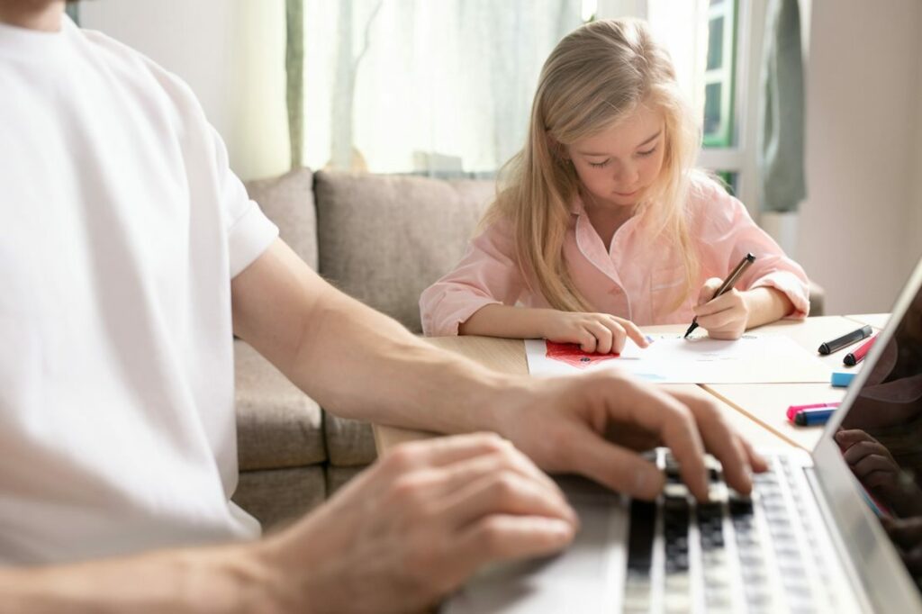 Un adulte tape au clavier à la droite d'une petite fille qui fait ses devoirs à la maison.