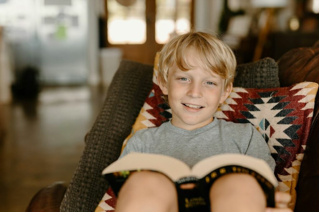 Un jeune élève blond, assis sur un canapé, est photographié en train de lire un livre.