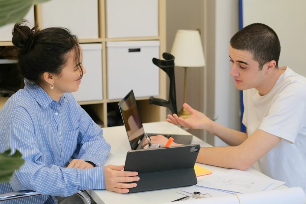 Deux adolescents travaillent autour d'une table, avec des feuilles, une tablette et un ordinateur.