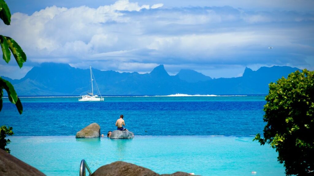 Vue d'un homme se tenant assis sur le bord d'une piscine, à côté de l'océan et les montagnes à l'horizon.