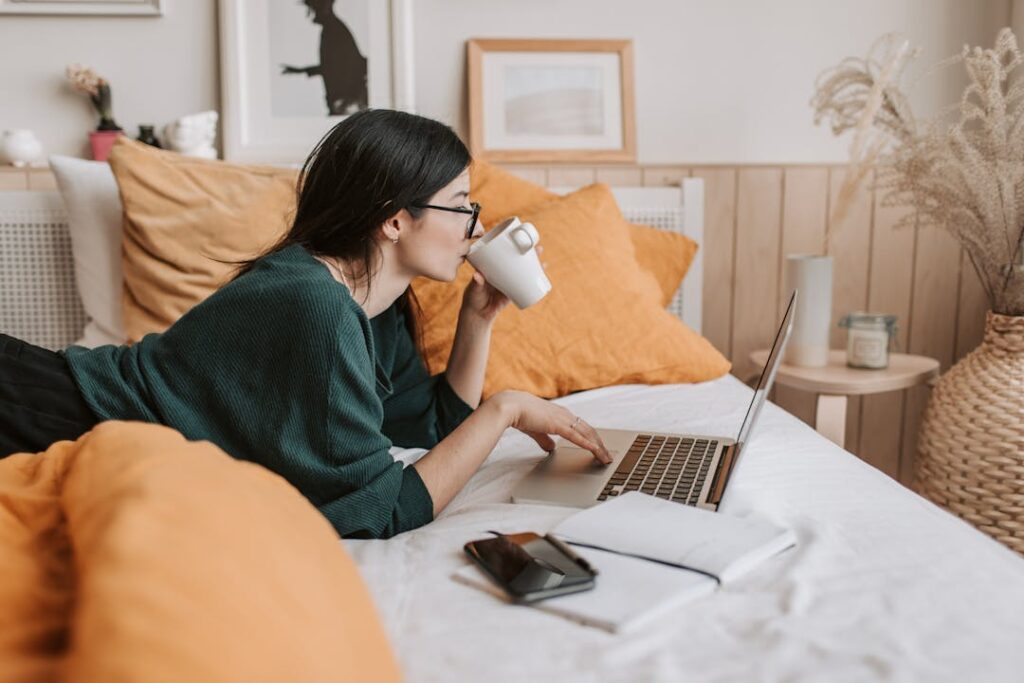 Une jeune femme allongée sur un lit en train de travailler sur son labtop, une tasse à la main gauche.