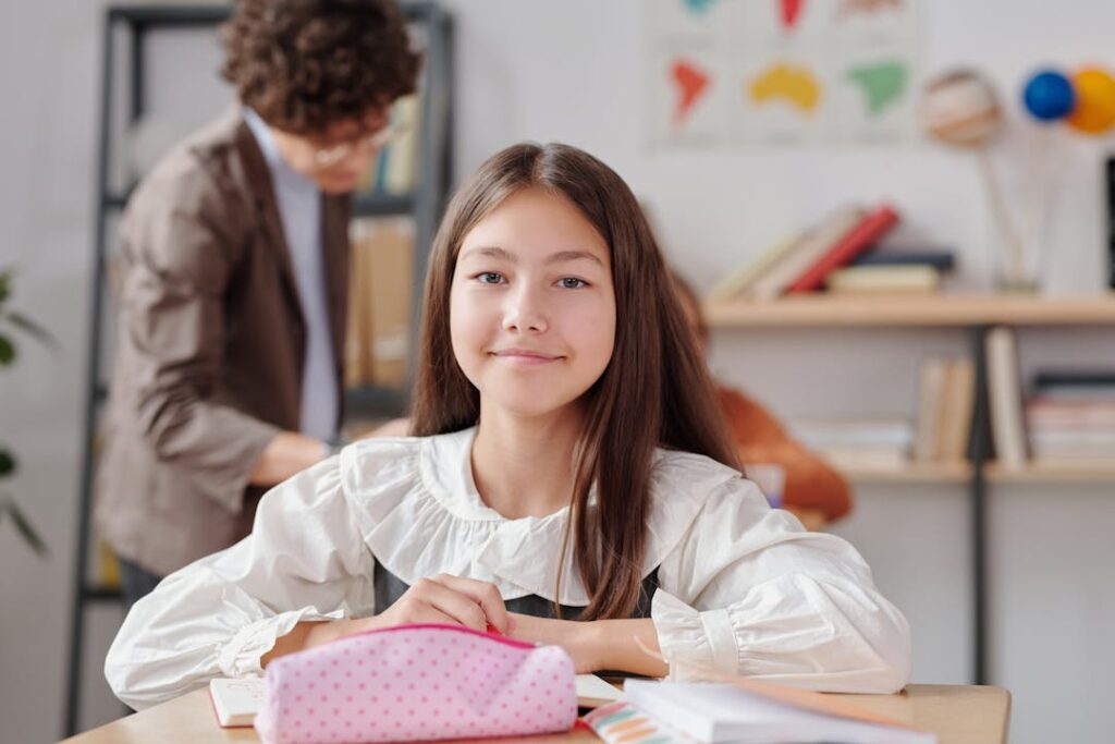 Une petite fille se tient droit à sa table d'école avec sa professeure derrière.