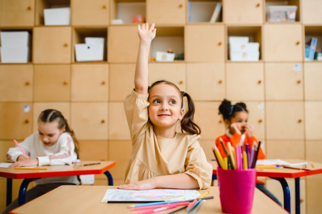 Une petite fille souriante lève la main en cours pour prendre la parole.