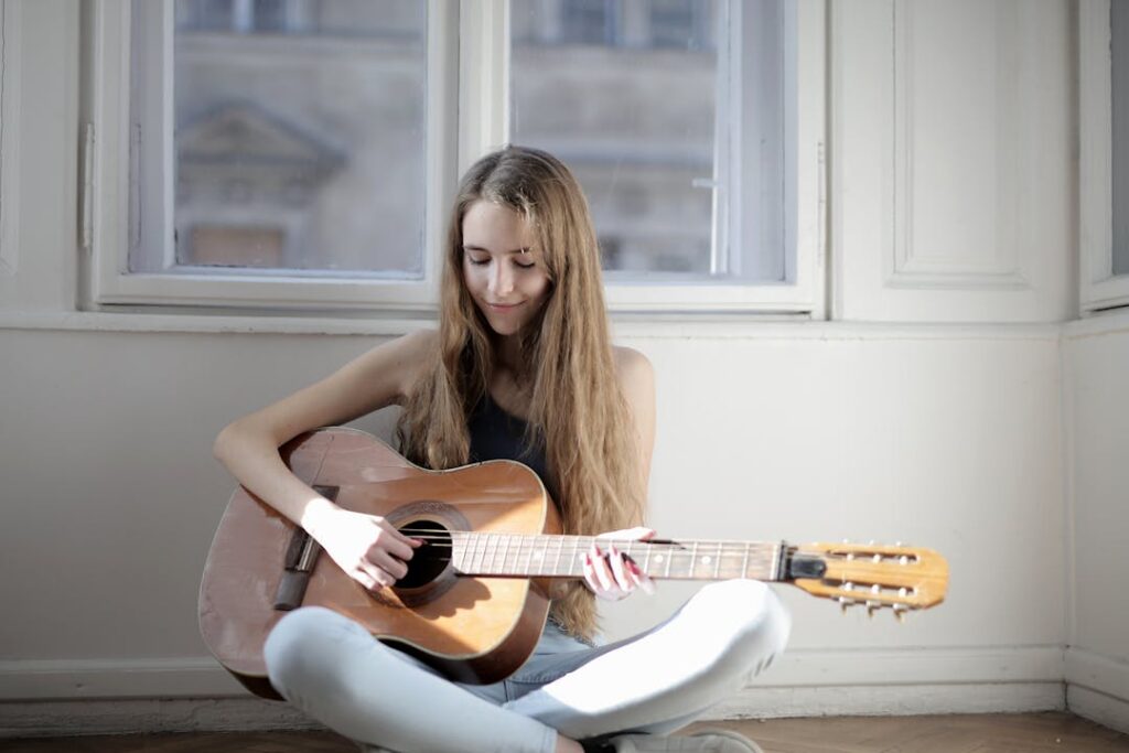 Une jeune fille joue de la guitare assise au sol, adossée à un mur blanc.