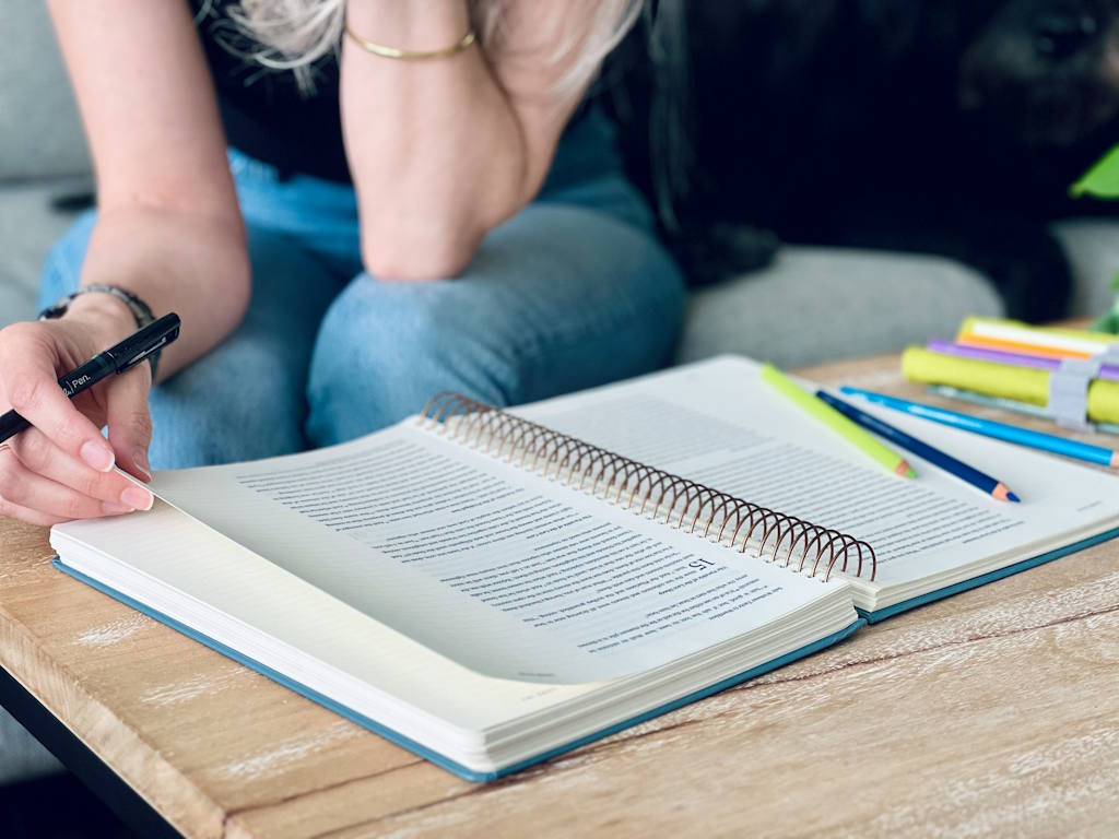 Une jeune femme est en train de lire un livre à reliure avec des crayons posés sur la table.