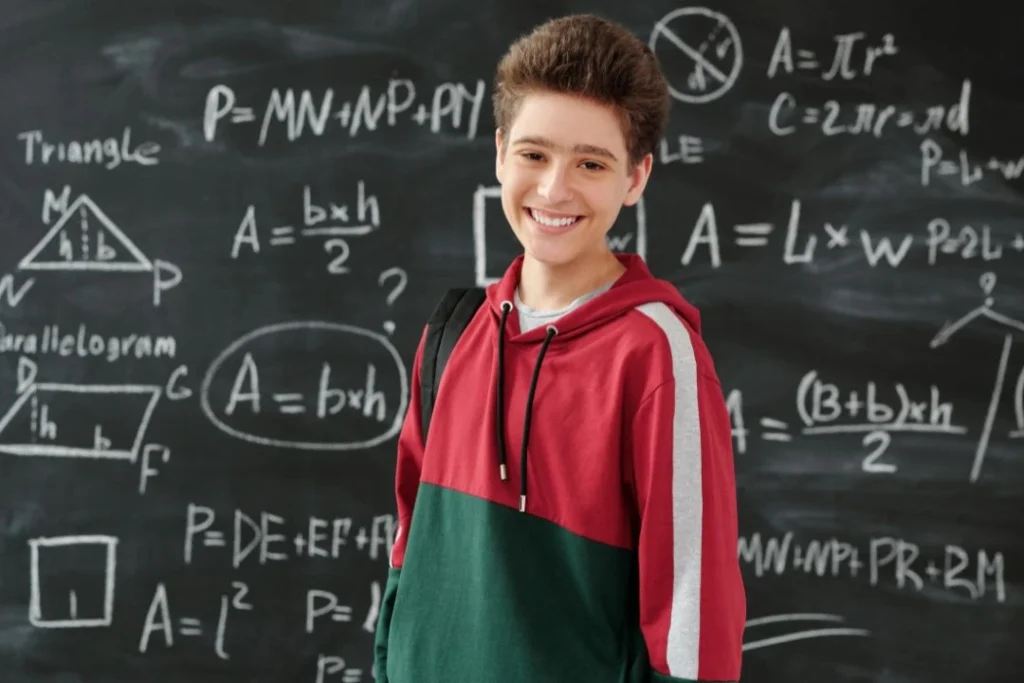Un jeune élève se tient debout, souriant devant un tableau noir rempli de formules mathématiques.