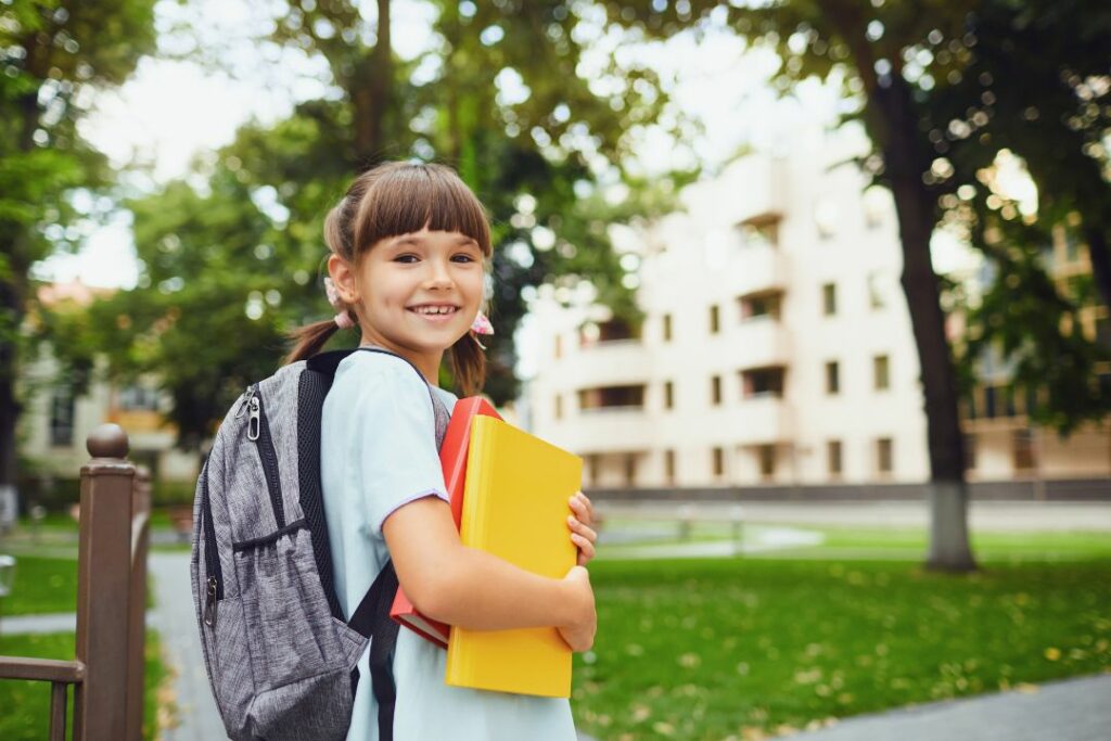 Une jeune enfant tient deux classeurs dans ses bras, près d'un parc arboré.
