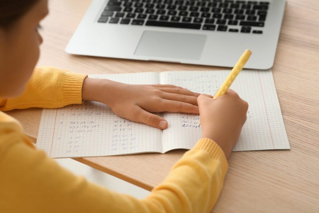 Élève résolvant des exercices de physique chimie sur un cahier avec un ordinateur portable ouvert à côté.